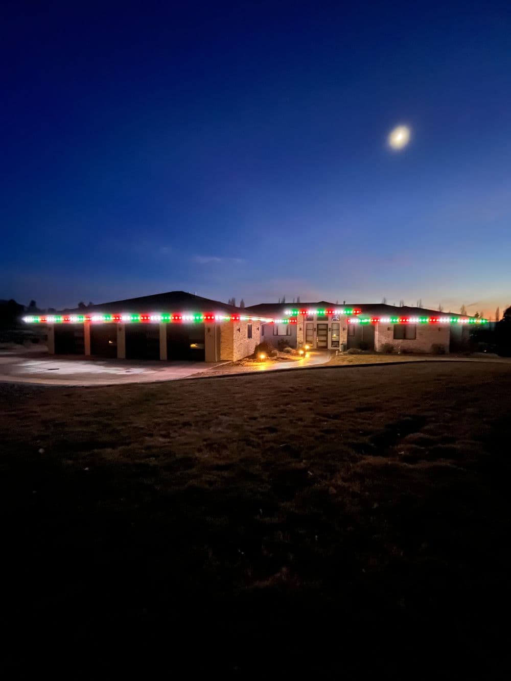 House decorated with colorful Christmas lights at dusk under a bright moon.